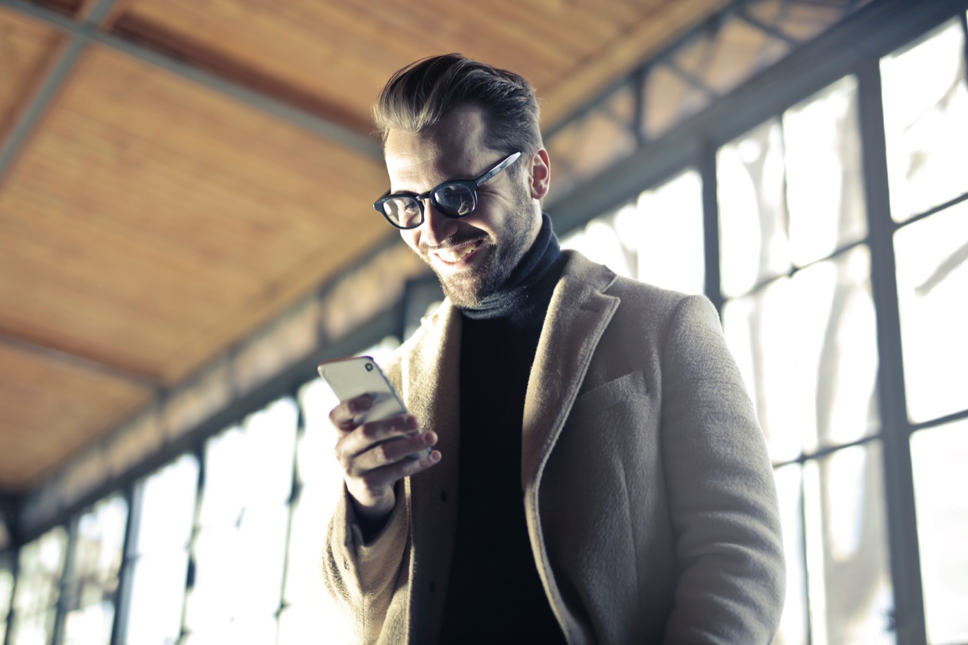 A well-dressed man in a wool coat and glasses smiling at his phone